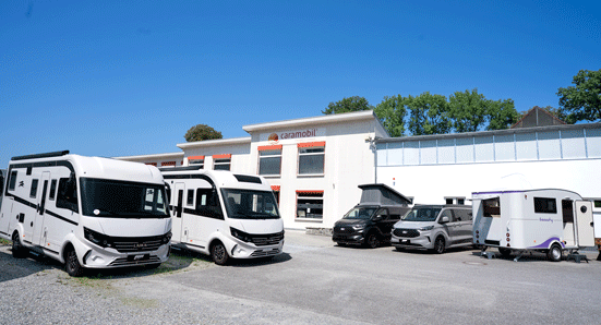Verschiedene weiße Wohnmobile und dunkle Campingbusse stehen vor dem hellen Caramobil Firmengebäude unter strahlend blauem Himmel.